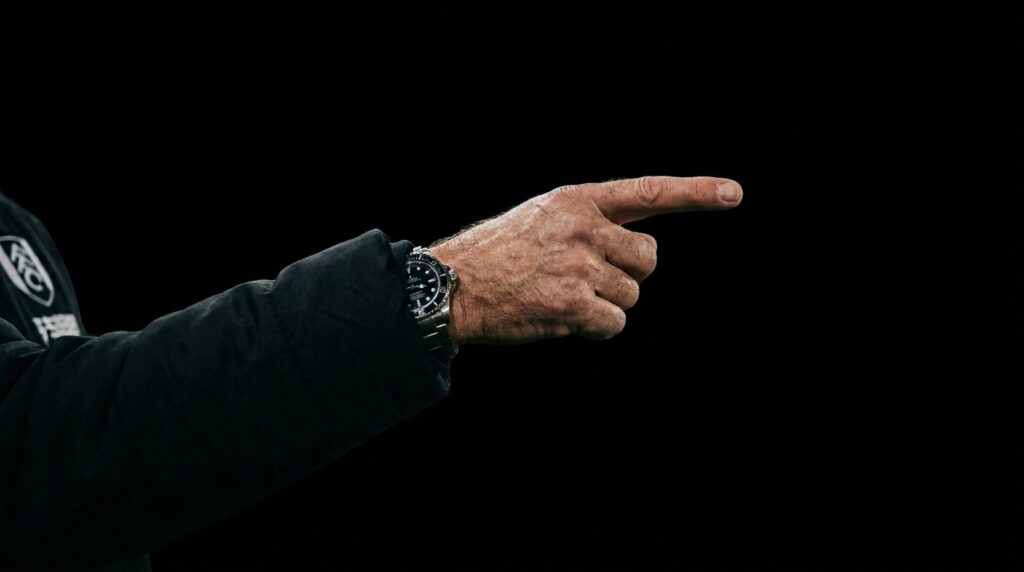 A close-up of a football manager's hand pointing aggressively from the technical area, isolated against a dark background.