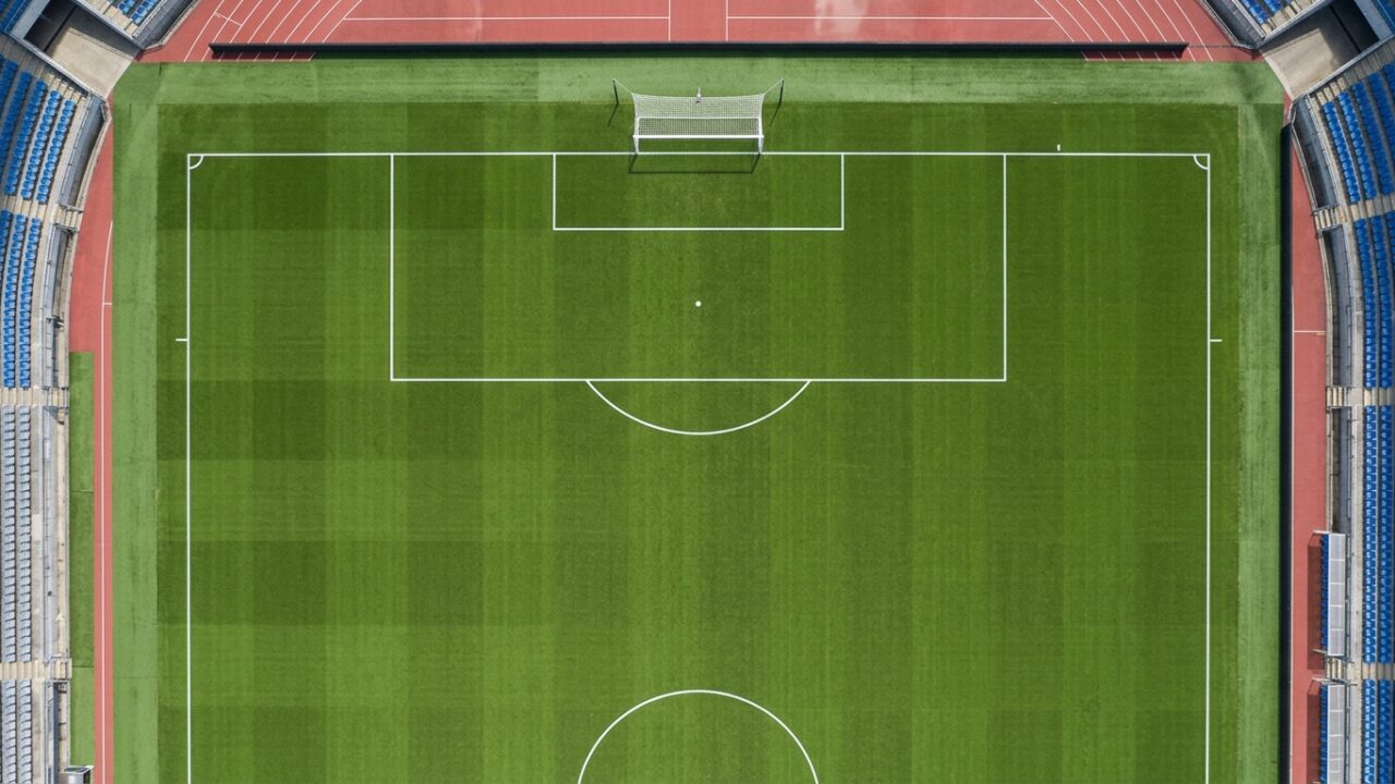A stark top-down view of a completely empty football pitch, emphasizing the geometric lines of the penalty area.